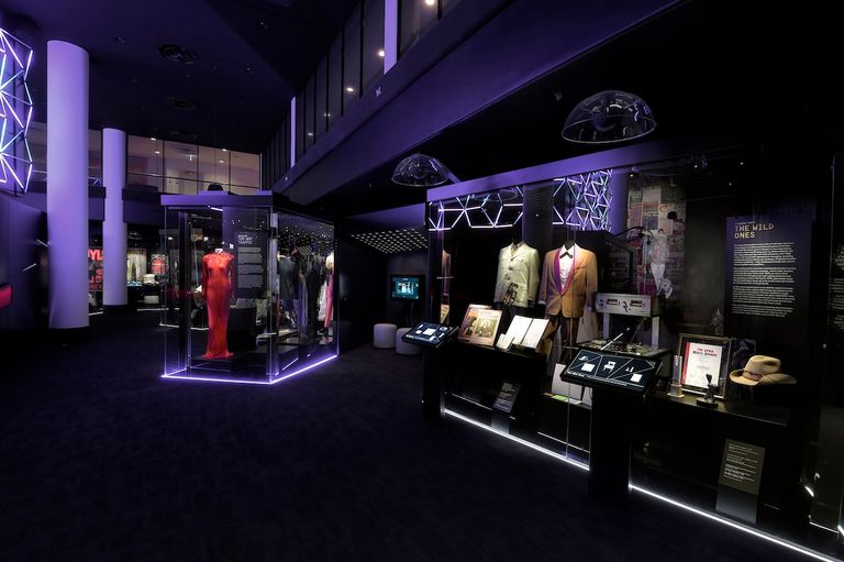 Alt Text: Australian Music Vault exhibition featuring illuminated display cases with iconic costumes, stage wear, awards, and music memorabilia under purple light.